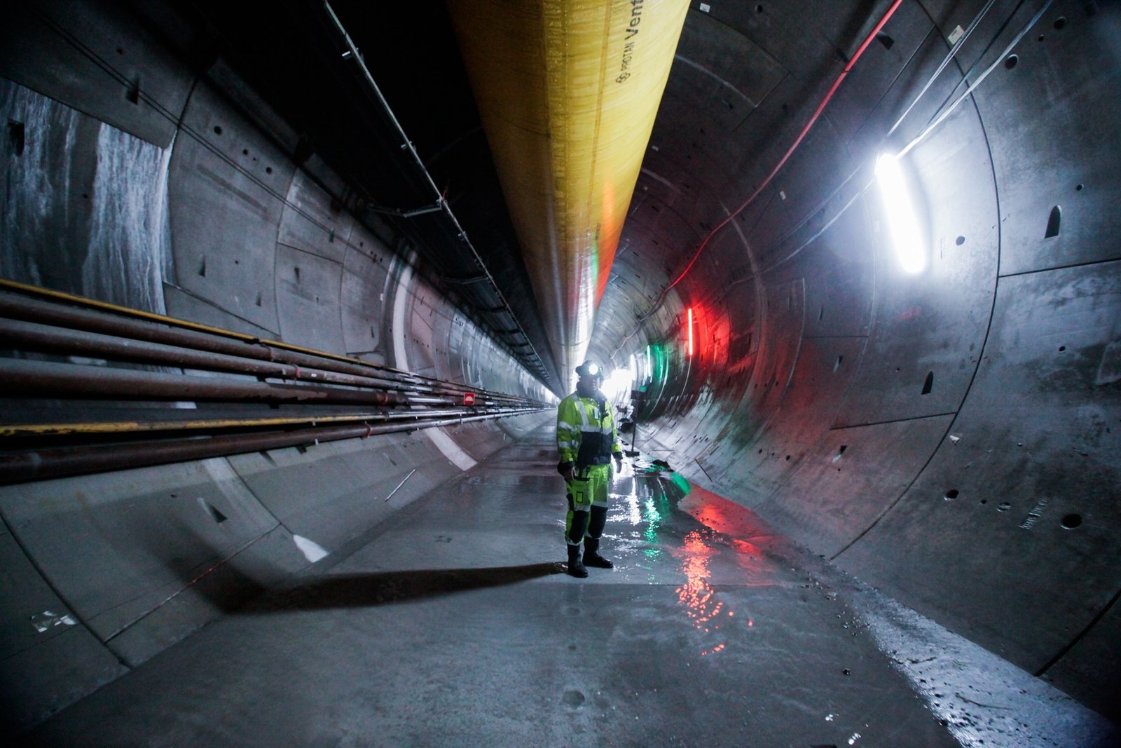 A worker in a hard hat and high-visibility clothing standing inside a long, lit concrete tunnel lined with pipes