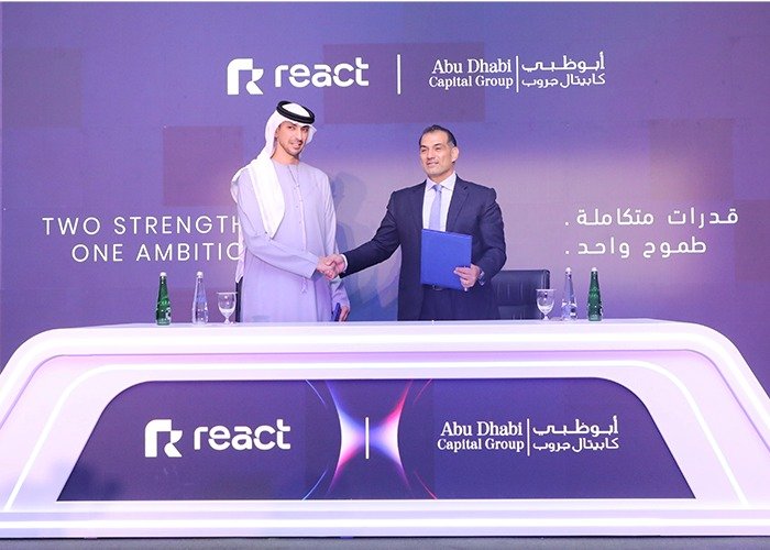 Two men shake hands behind a signing desk carrying React and Abu Dhabi Capital Group logos