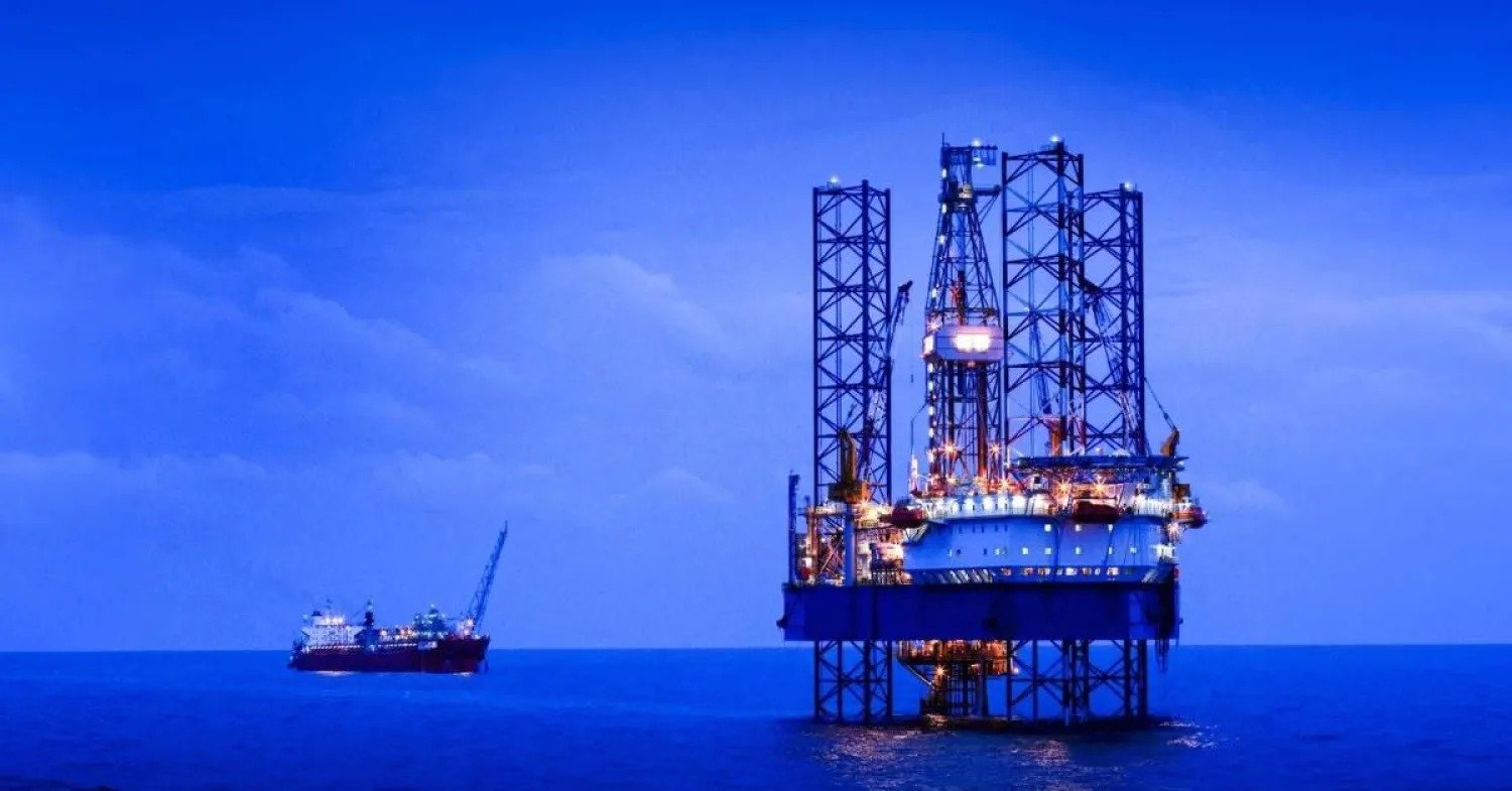 An ADES Holding offshore jackup drilling rig lit at dusk at sea, with a support vessel in the background