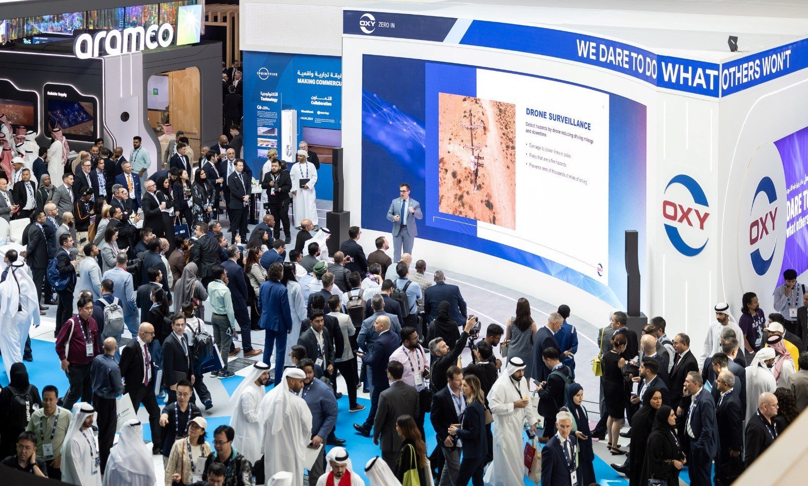Crowds of visitors walk between exhibition stands, including Aramco and OXY booths, as a presenter speaks in front of a large screen at ADIPEC