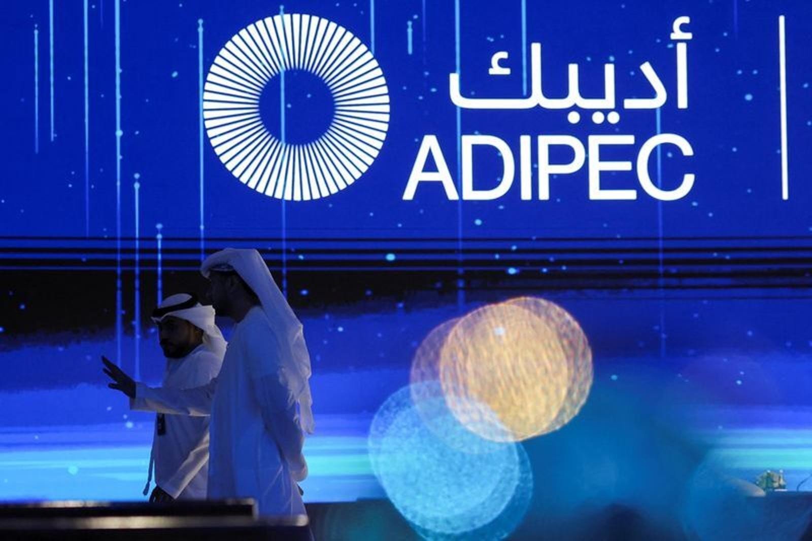 Two men in traditional Emirati dress walk past a large ADIPEC sign on a stage in Abu Dhabi