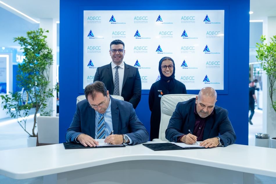 Two men sign documents at a white table as a man and a woman stand behind them in front of a branded backdrop