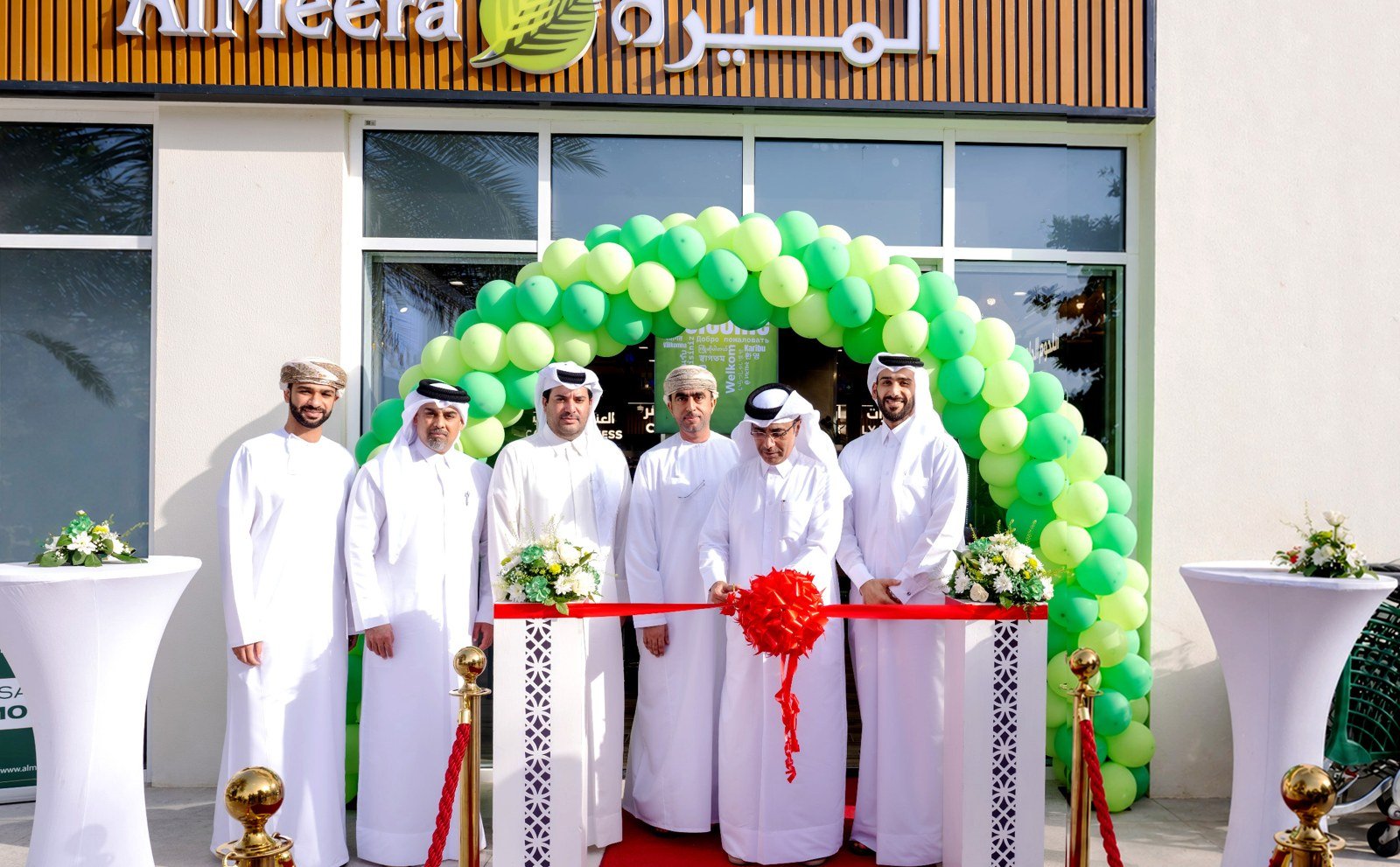Men in traditional Qatari dress stand at a ribbon-cutting outside a new Al Meera supermarket decorated with green balloons