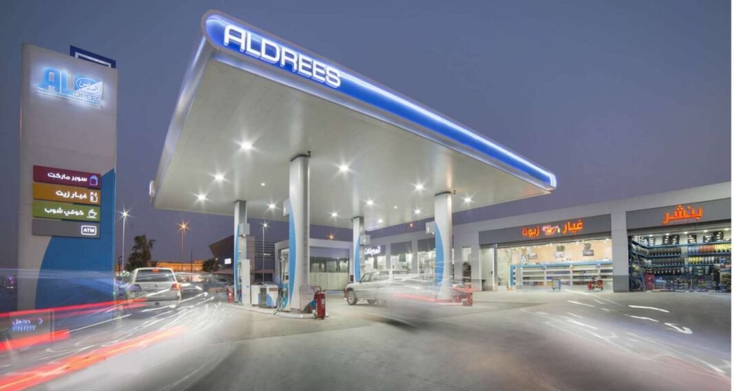 An Aldrees fuel service station at dusk with a lit canopy and shops