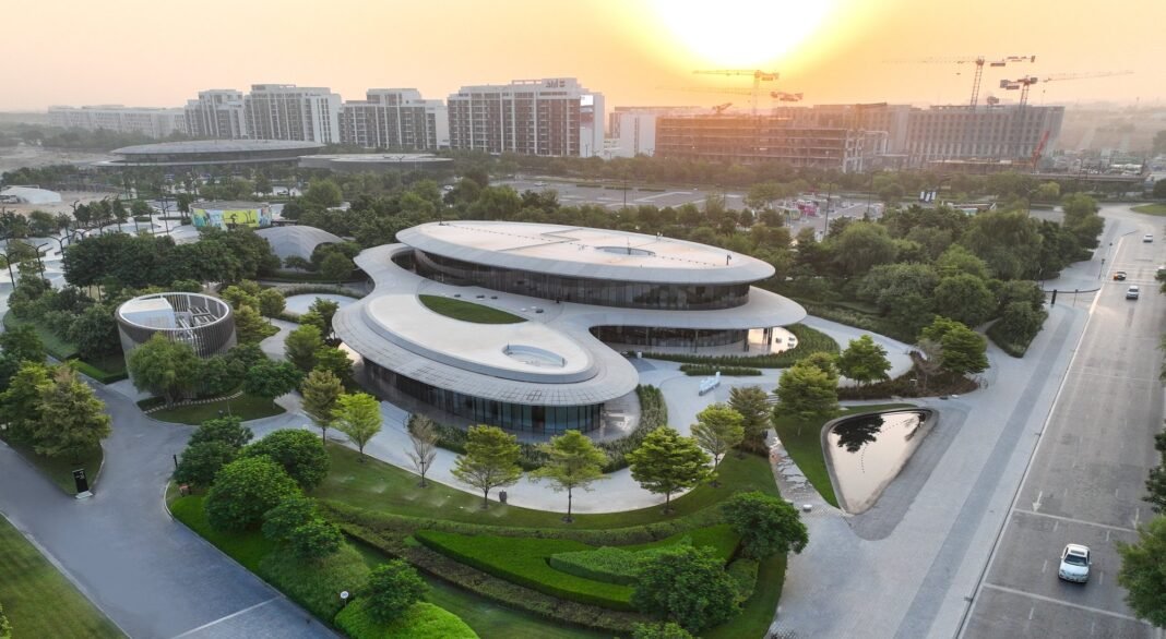 Aerial view at sunset of a curved, low-rise building surrounded by landscaped gardens, with apartment blocks and construction cranes behind it