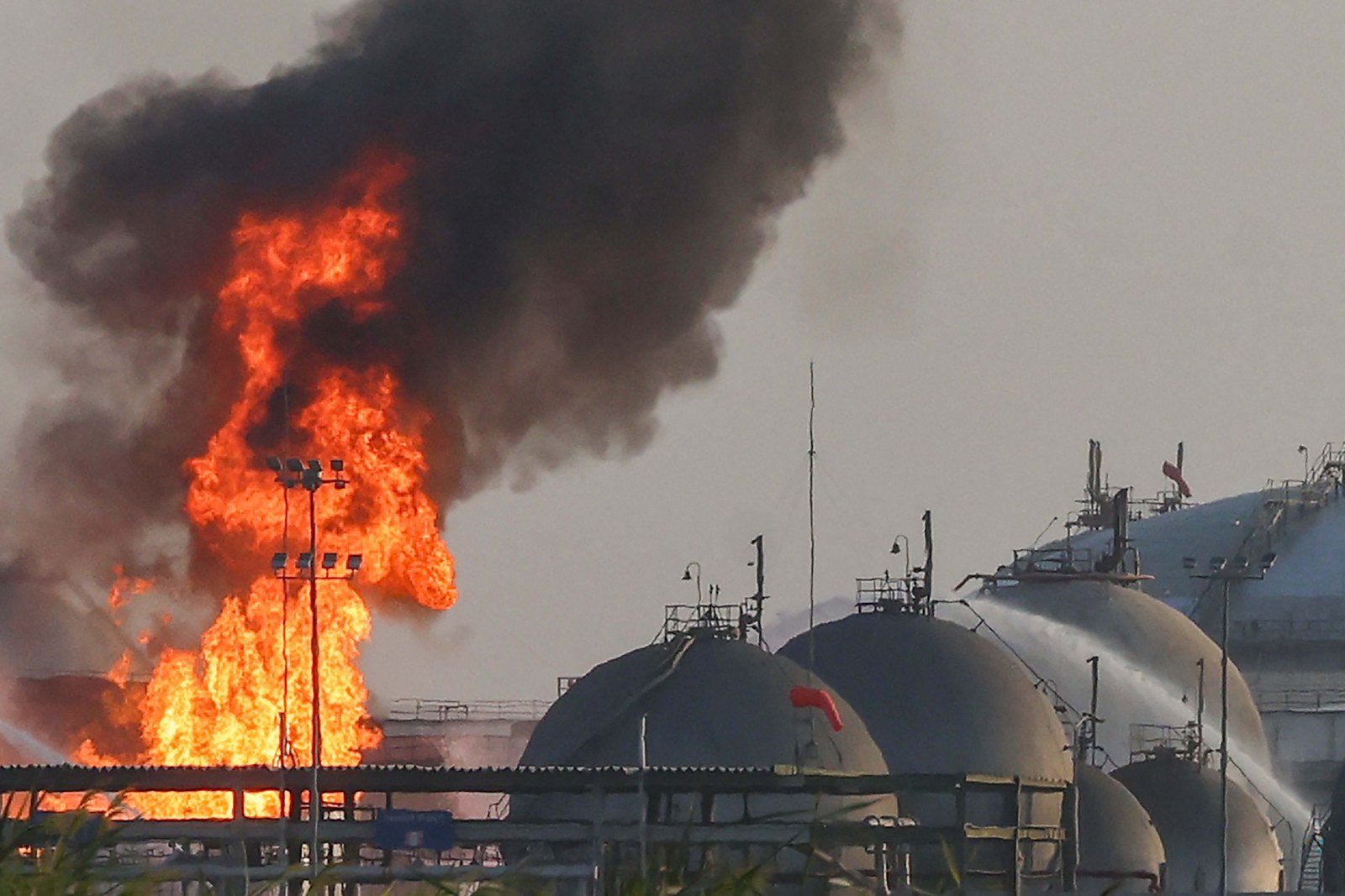 Flames and black smoke rise beside spherical storage tanks at an Aramco facility as water is sprayed on the tanks