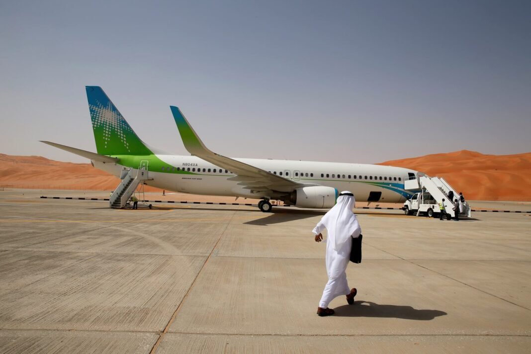 A man in a white thobe and headdress walks across an airfield toward a white and green Aramco passenger jet, with orange sand dunes behind