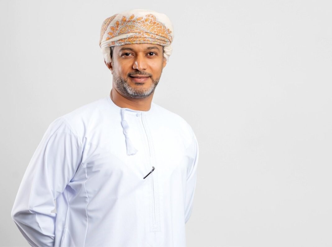 Portrait of a smiling man in a white Omani dishdasha and an embroidered cream and orange turban against a light gray background