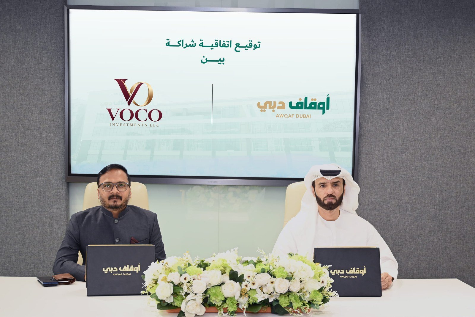 Two men seated at a table with white flowers sign documents in front of a screen showing the Voco Investments and Awqaf Dubai logos