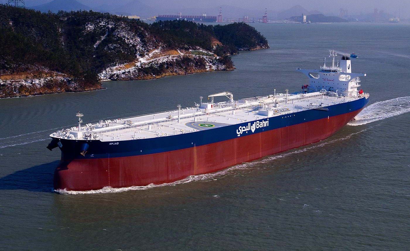 A Bahri very large crude carrier with a red hull and the company name on its side sailing near a rocky coastline