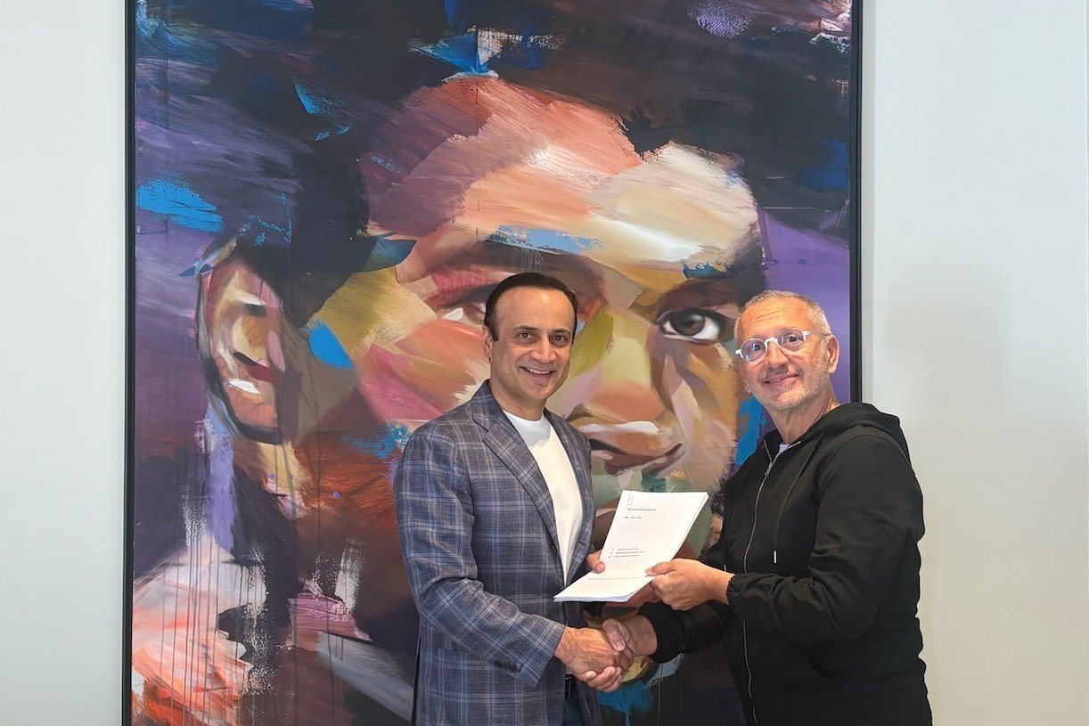 Sanjeev Nanda and Christophe Caucino shake hands holding a document in front of a large colorful portrait painting