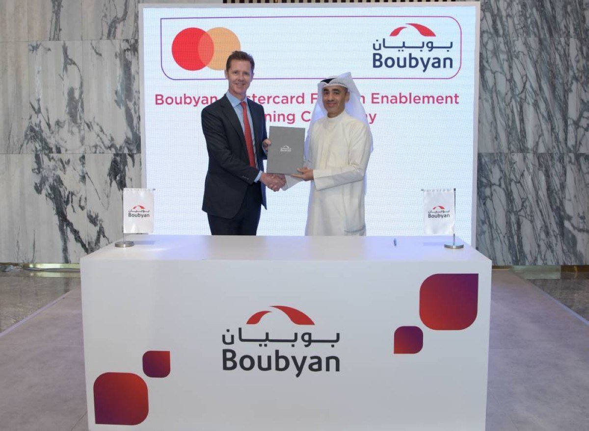 Two men shake hands and hold a document beside a podium in front of a Boubyan and Mastercard backdrop