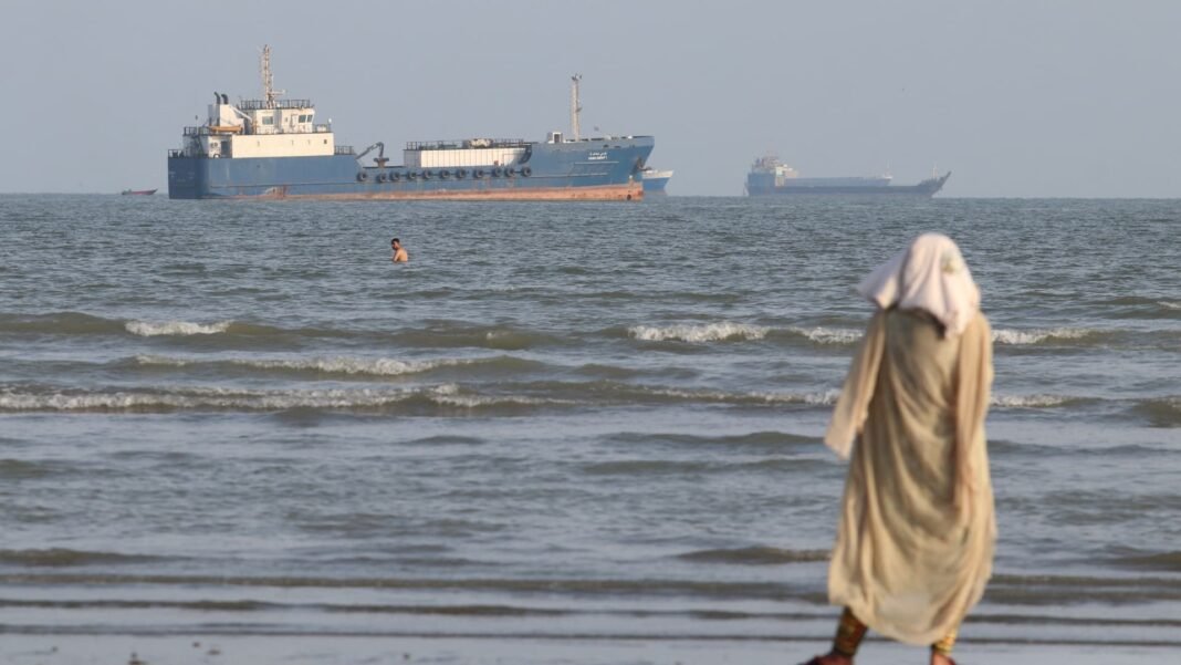 A woman in a headscarf walks along a beach as cargo vessels sit offshore in the Strait of Hormuz
