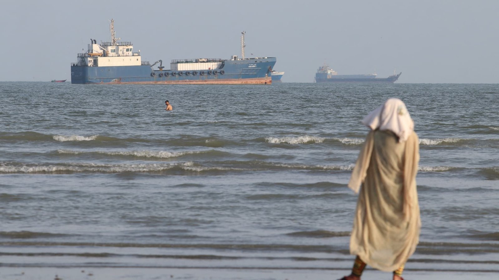 A woman in a headscarf walks along a beach as cargo vessels sit offshore in the Strait of Hormuz