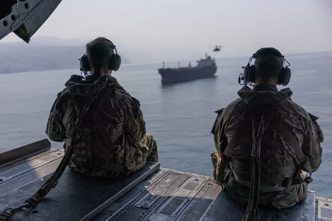 Two US military officers sit at the open rear ramp of a CH-47 Chinook helicopter watching a tanker sail through the Strait of Hormuz