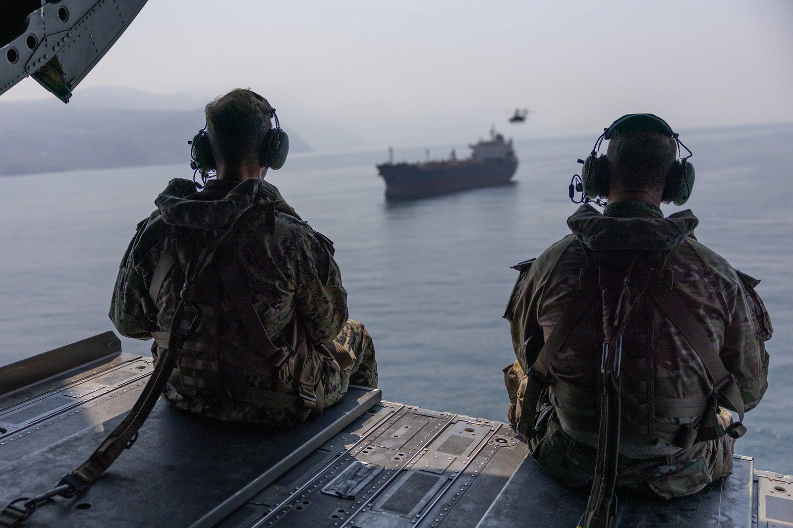 Two US military officers sit at the open rear ramp of a CH-47 Chinook helicopter watching a tanker sail through the Strait of Hormuz