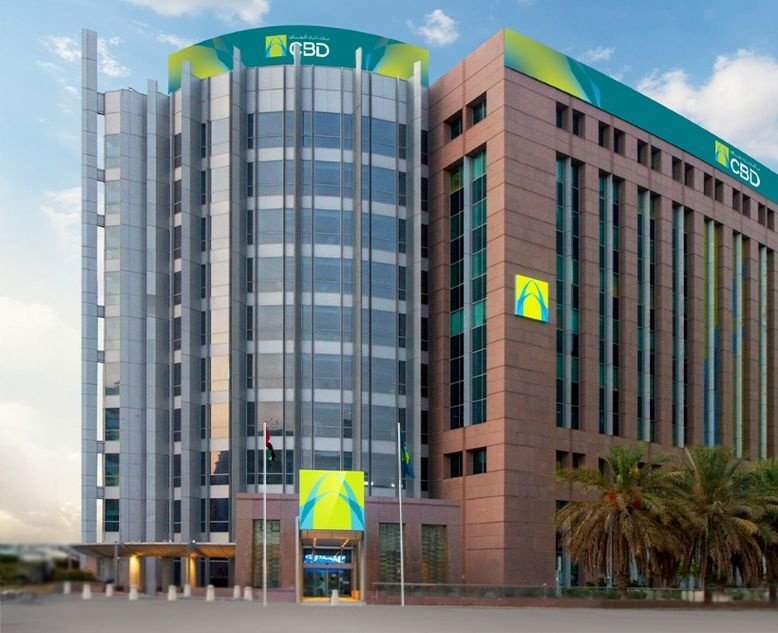A multi-story office building with a curved glass tower and a stone-clad wing bearing the CBD logo, with palm trees at its entrance