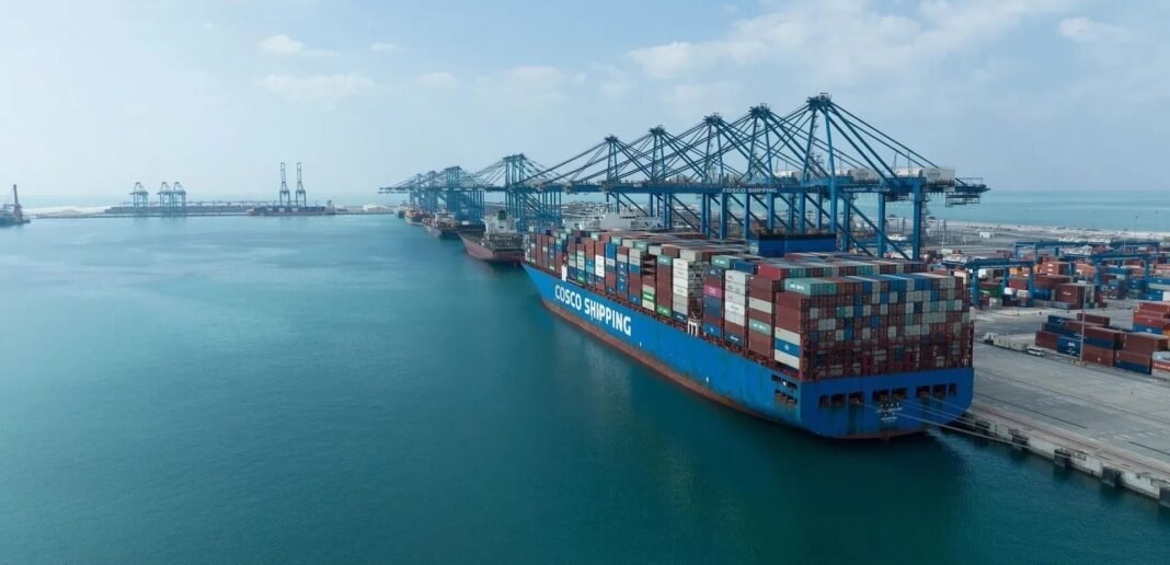 A large COSCO Shipping container vessel loaded with containers berthed beneath blue ship-to-shore cranes