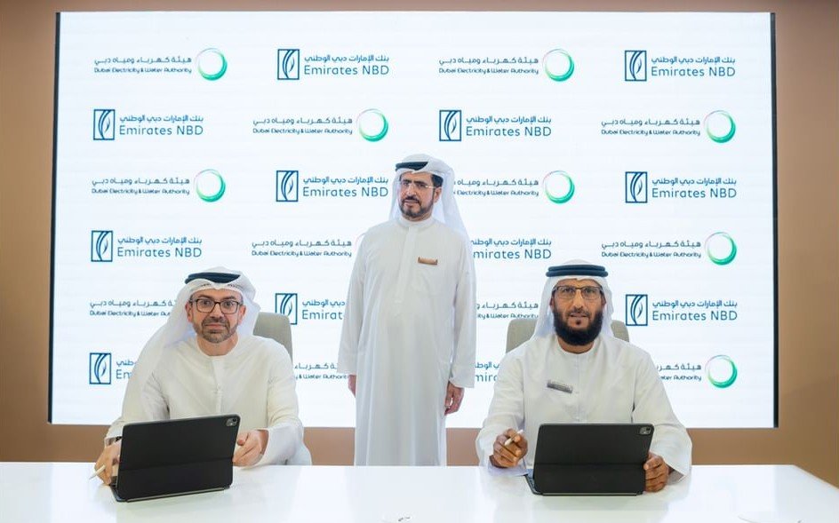 A man in a white kandura stands between two seated officials signing documents on tablets in front of a backdrop with DEWA and Emirates NBD logos