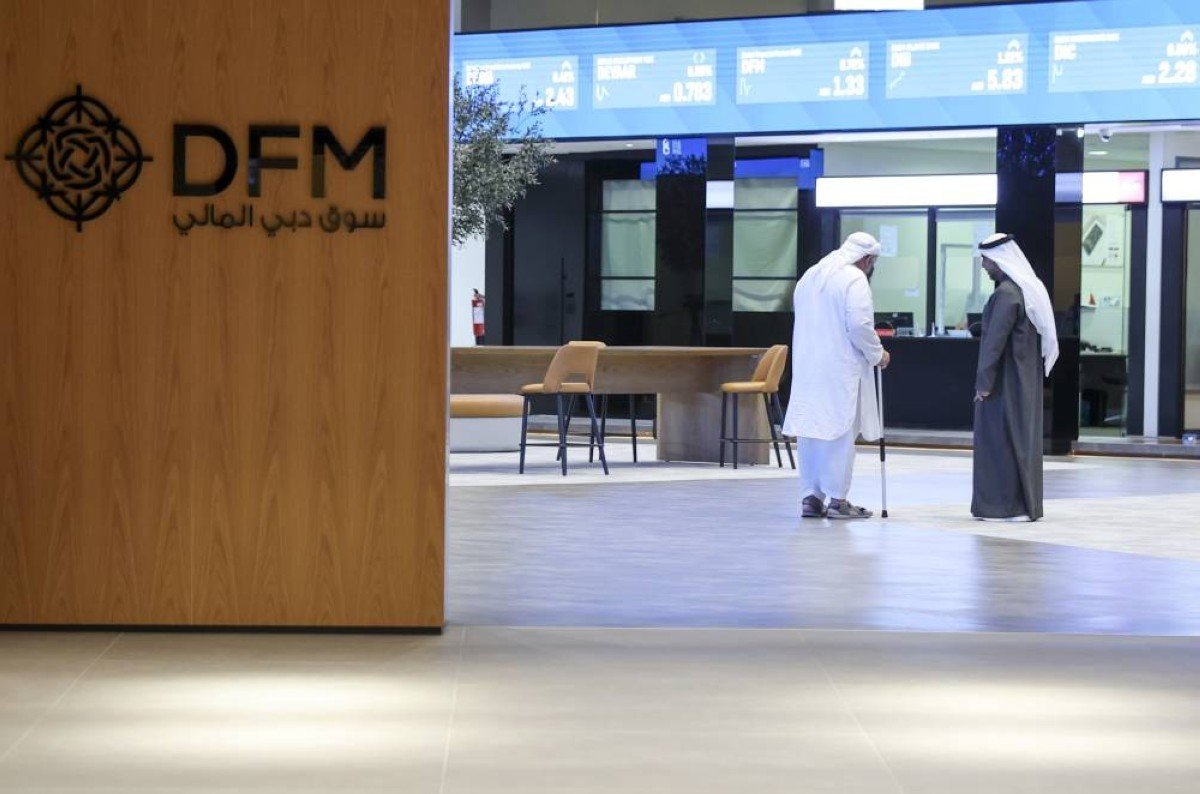 Two men talk in the lobby of the Dubai Financial Market beside the DFM sign, with price screens overhead
