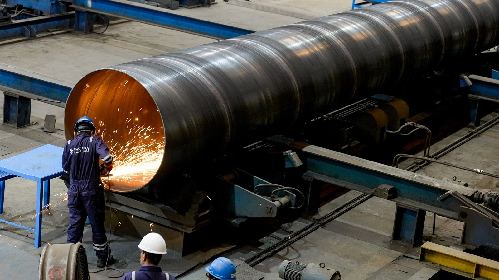 A worker in overalls grinds the inside edge of a large spiral-welded steel pipe as sparks fly in a factory hall