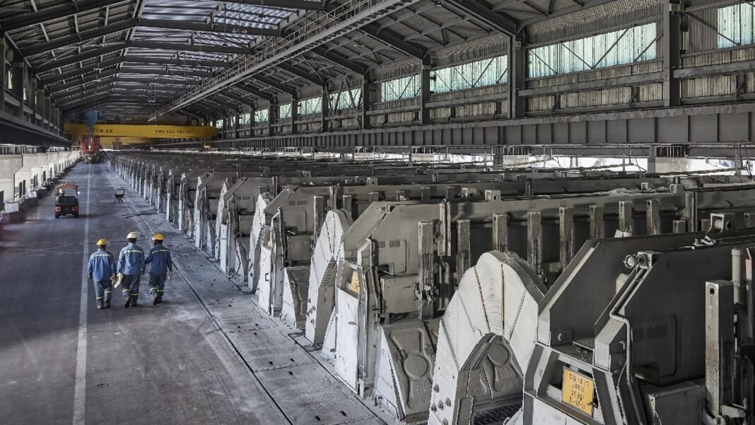 Three workers in blue overalls and hard hats walk beside a long row of aluminium reduction cells inside the Al Taweelah smelter potline