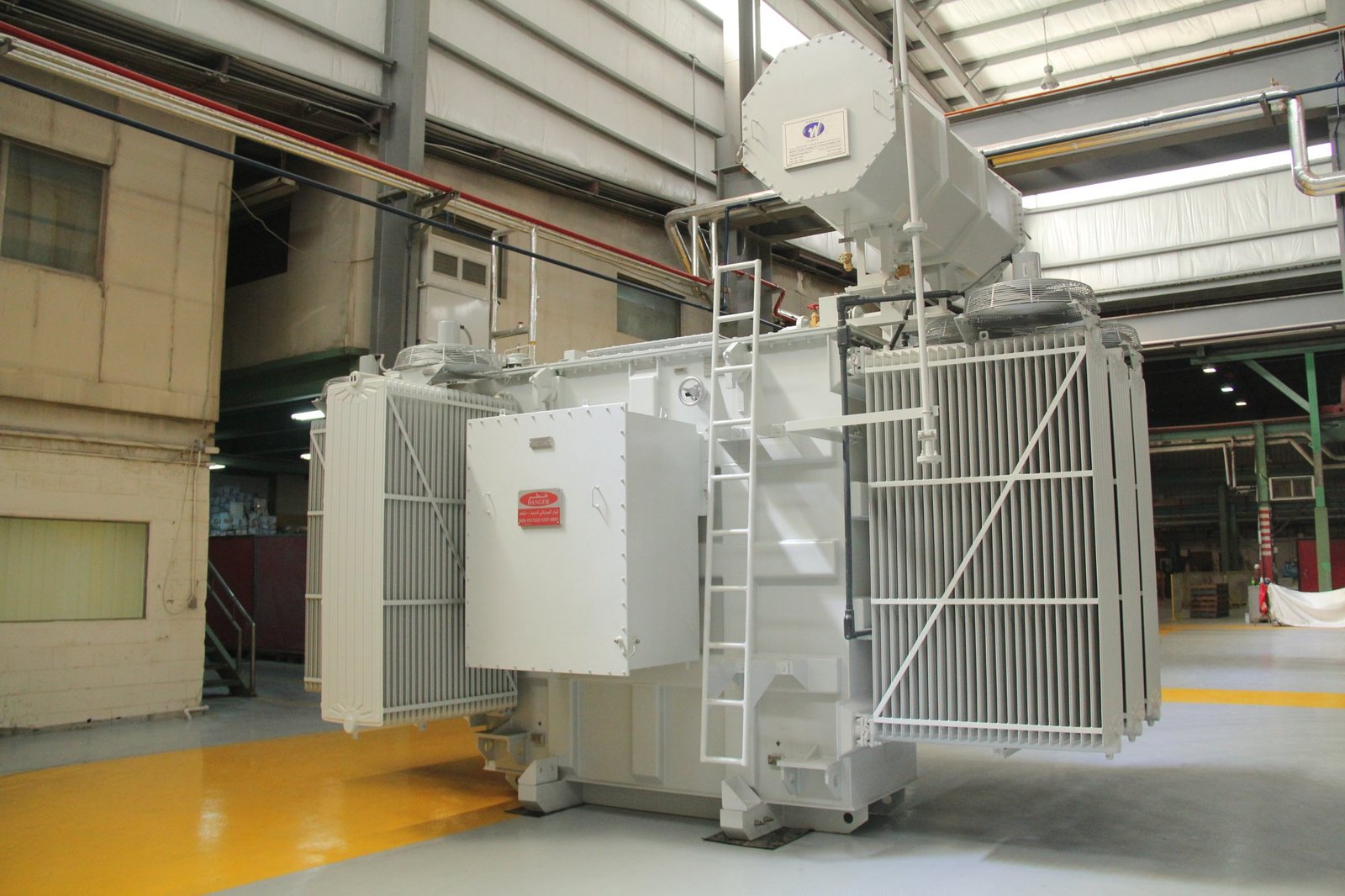 A large grey power transformer with cooling fins and a ladder stands on the floor of a factory hall