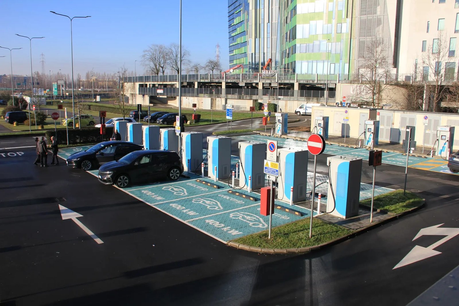 A row of electric vehicle charging stations with cars parked in marked bays outside office buildings