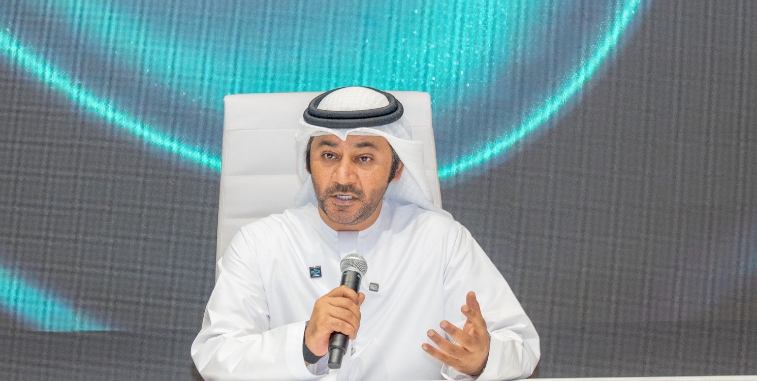 An Emirati executive in a white kandura speaks into a microphone at a desk during a ceremony at the Abu Dhabi Securities Exchange