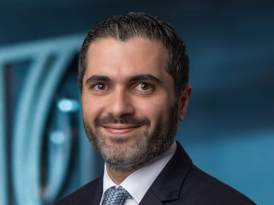 Portrait of a smiling bearded man in a dark suit and blue patterned tie against a blue background