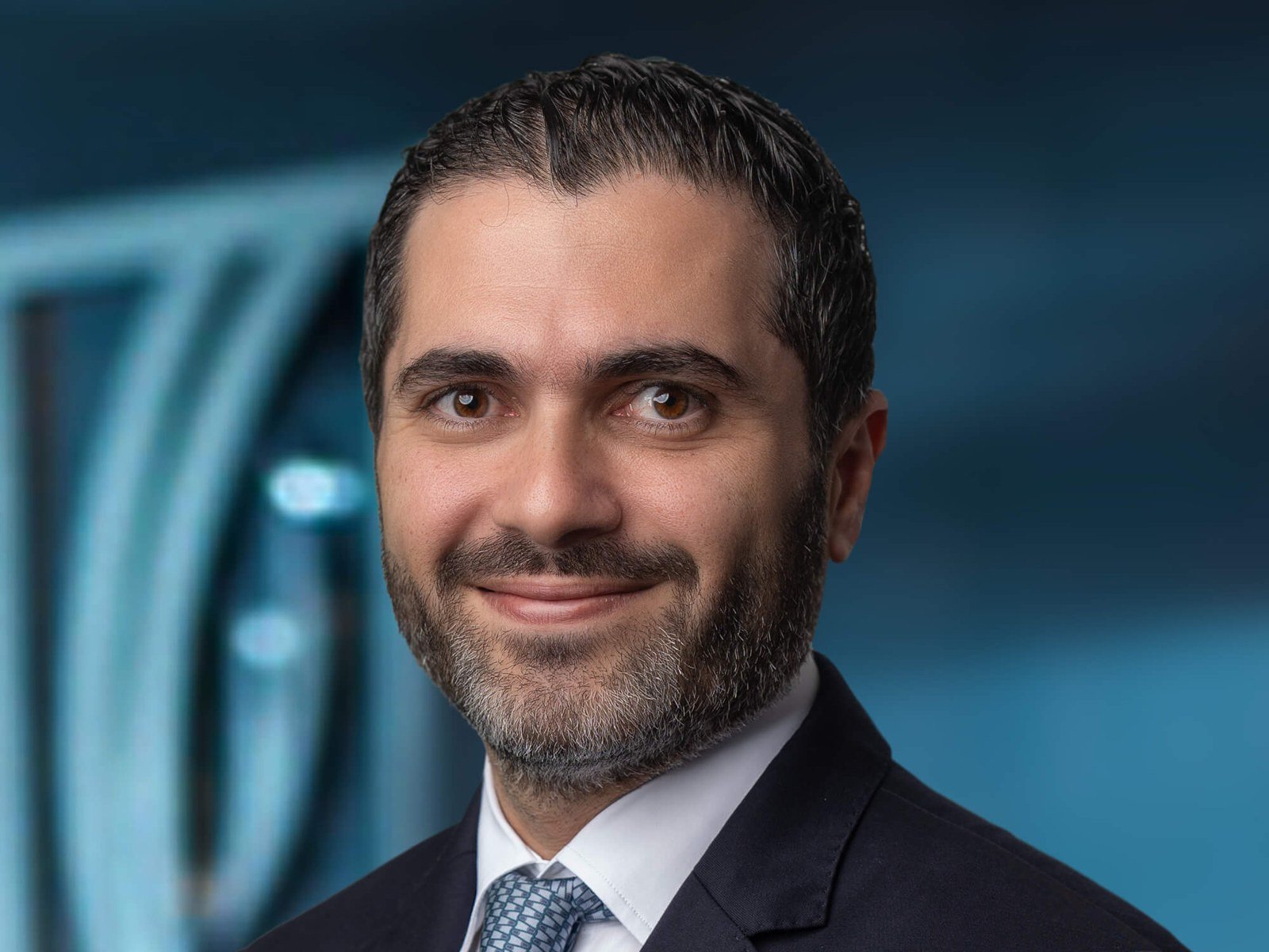 Portrait of a smiling bearded man in a dark suit and blue patterned tie against a blue background