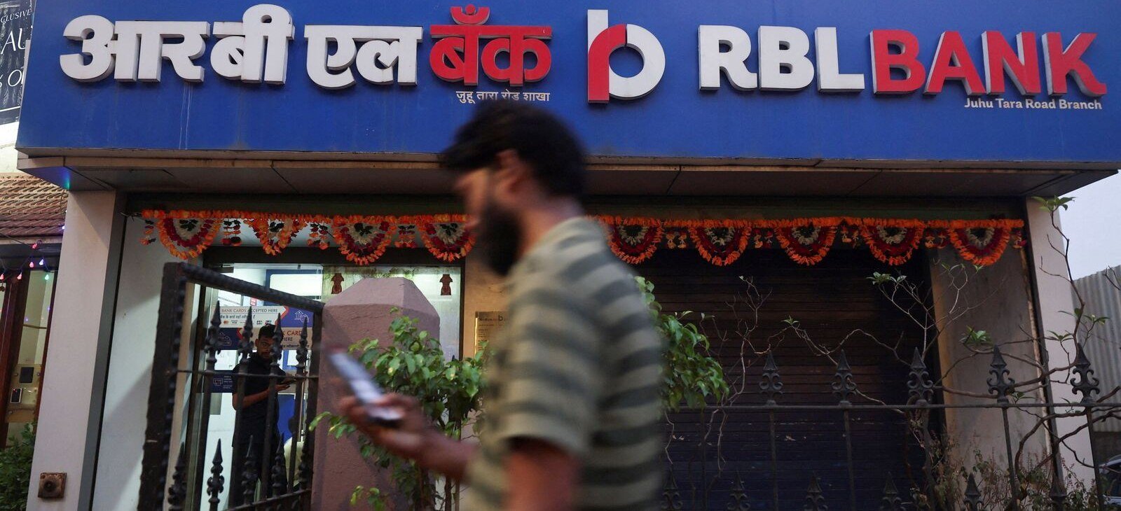 A man looking at his phone walks past an RBL Bank branch with a blue sign and flower garlands over the entrance