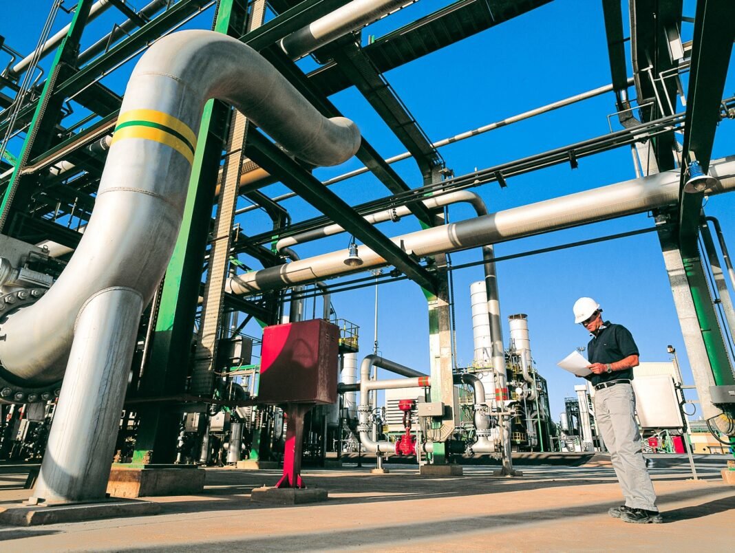 A worker in a white hard hat reads a document beside large silver pipes at a gas processing plant under a blue sky