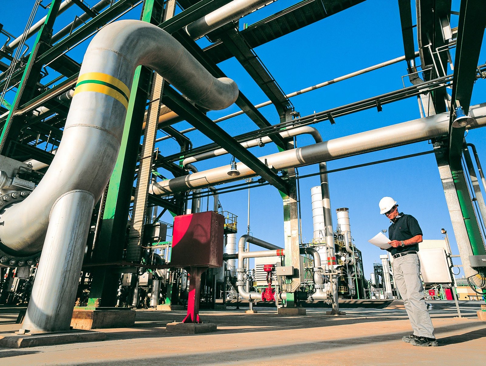 A worker in a white hard hat reads a document beside large silver pipes at a gas processing plant under a blue sky