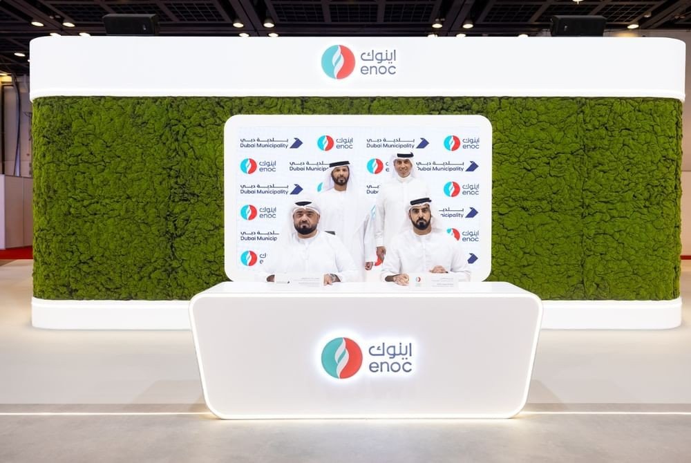 Officials in white kanduras seated and standing at a signing desk in front of a backdrop showing ENOC and Dubai Municipality logos
