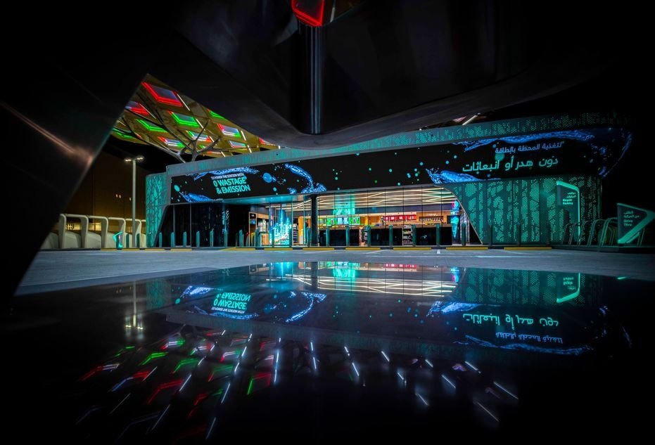 An ENOC service station lit at night with a sign reading zero wastage and emission, reflected in a wet forecourt