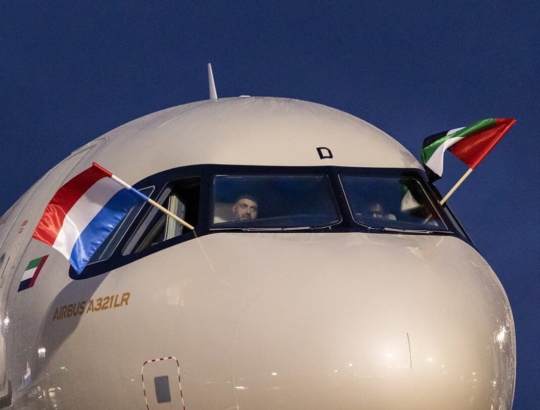 The nose of a white Airbus A321LR with a Luxembourg flag and a UAE flag flying from its cockpit windows against a dark blue sky