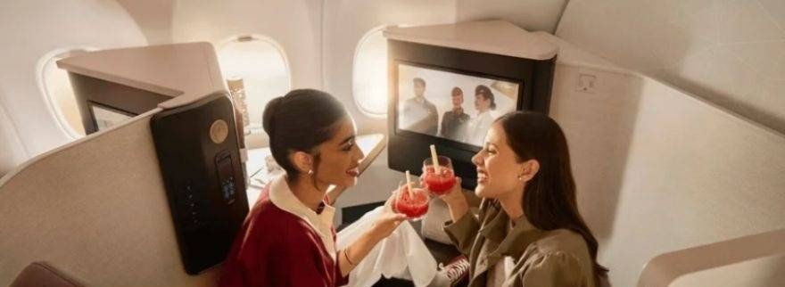 Two women smile and raise red drinks in adjacent aircraft seats in front of a seatback screen