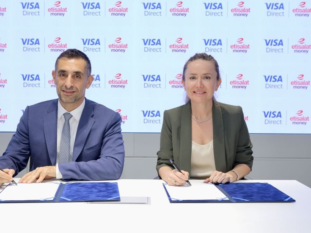A man and a woman sign documents at a table in front of a screen showing Visa Direct and etisalat money logos