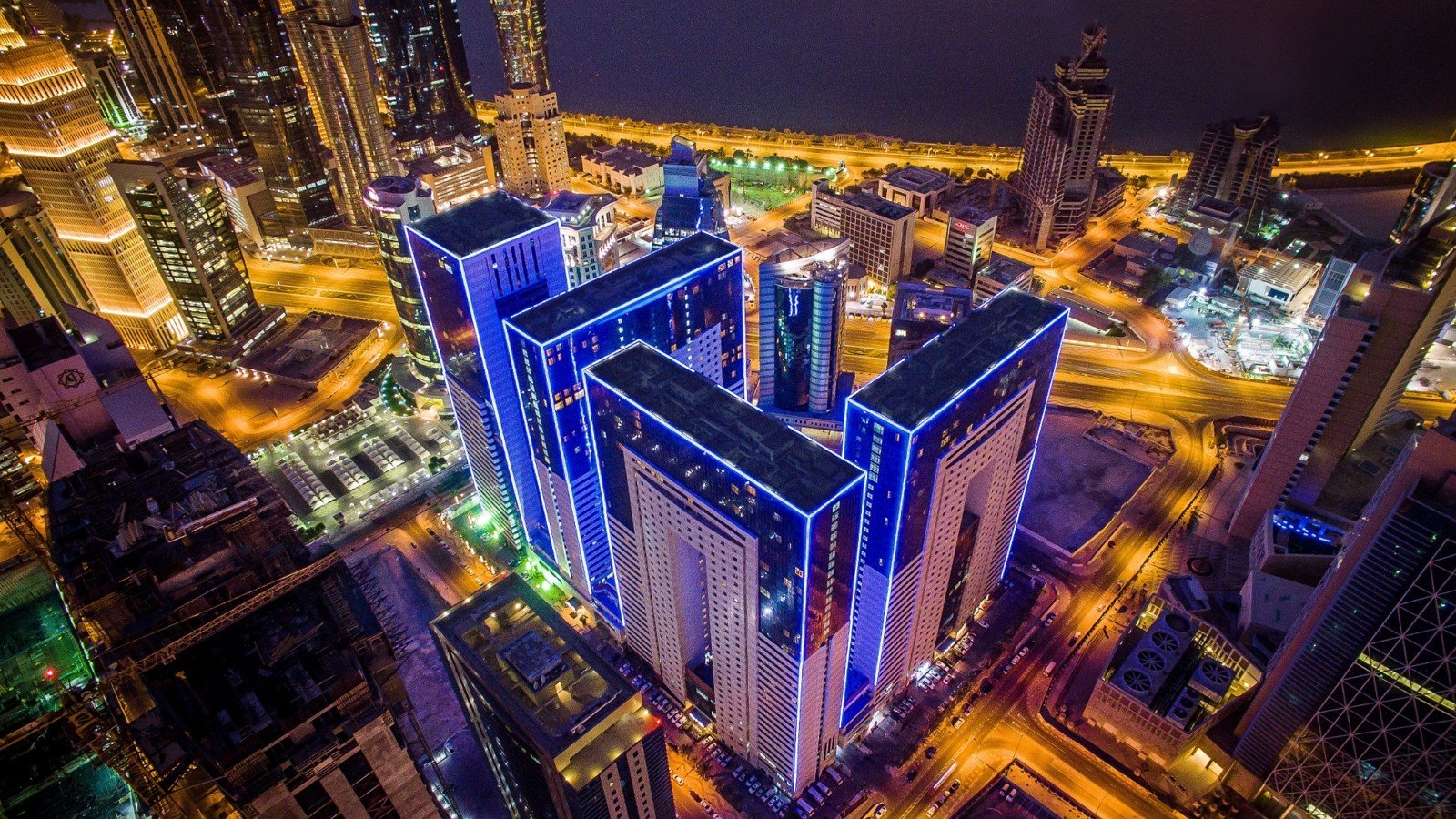 An aerial night view of four high-rise towers outlined in blue light among lit roads and buildings, with the sea in the background