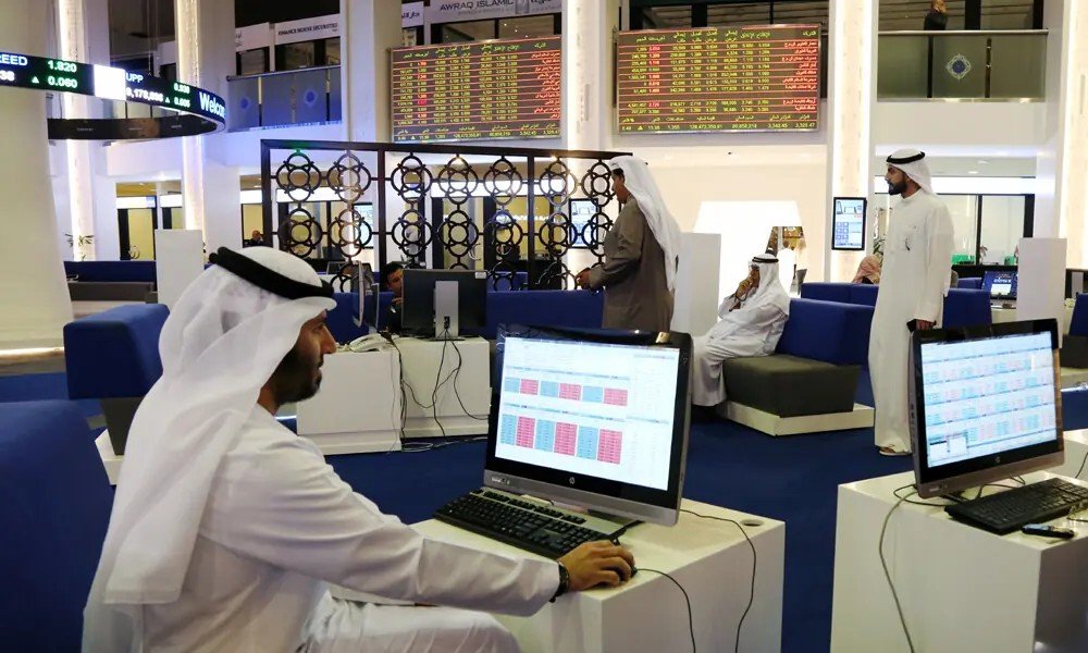 Traders in white kanduras work at computer terminals on the Dubai Financial Market trading floor beneath electronic share price boards