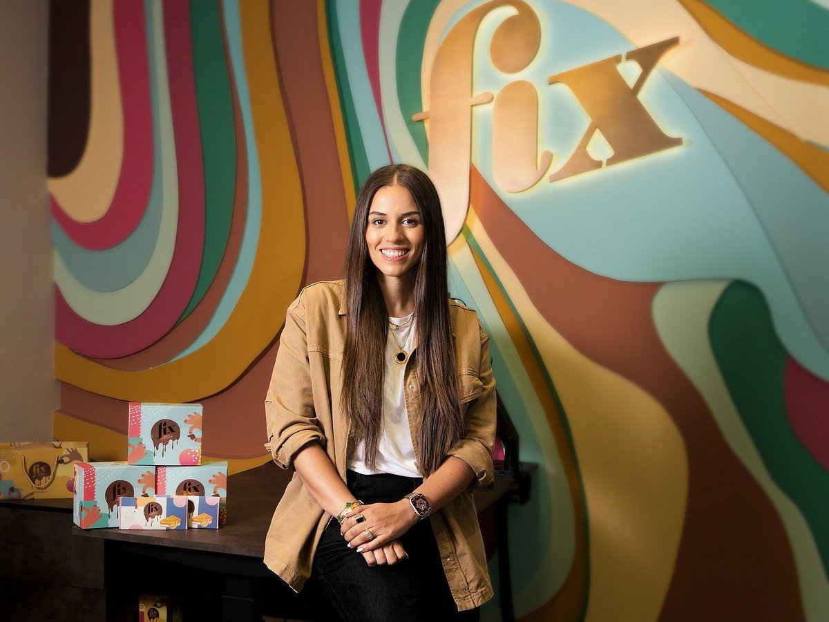 FIX Dessert Chocolatier co-founder Sarah Hamouda in front of a wall bearing the FIX logo, with boxes of chocolate on a desk beside her