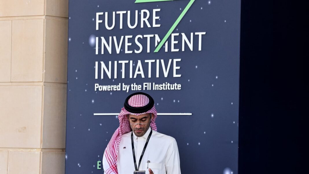 A man in a red and white shemagh checks his phone in front of a Future Investment Initiative sign in Riyadh