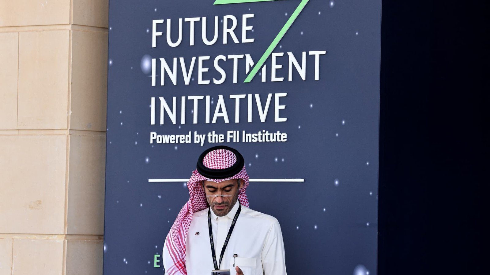A man in a red and white shemagh checks his phone in front of a Future Investment Initiative sign in Riyadh