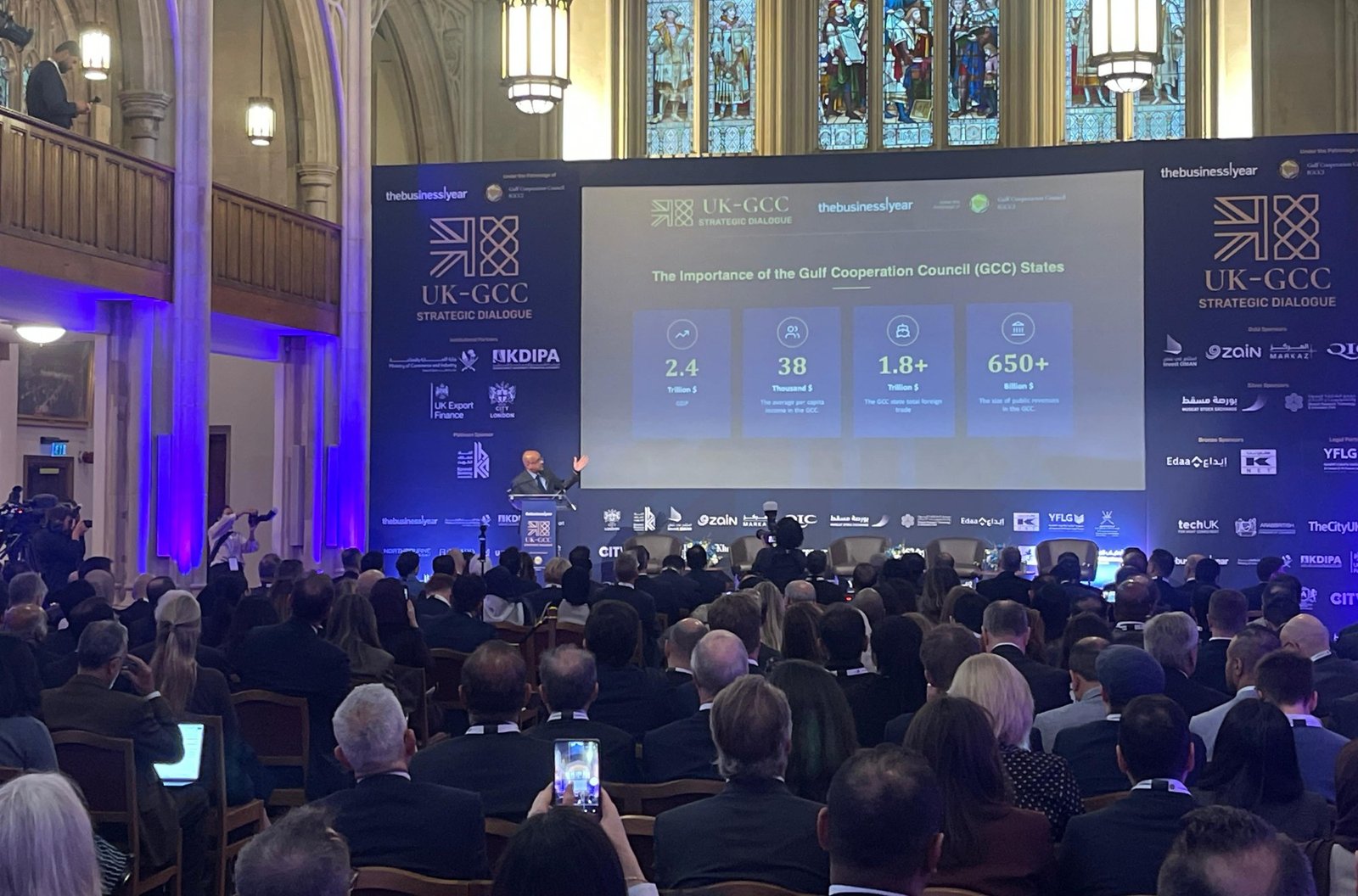 A speaker addresses a packed hall with stained-glass windows in front of a large screen showing UK-GCC Strategic Dialogue figures