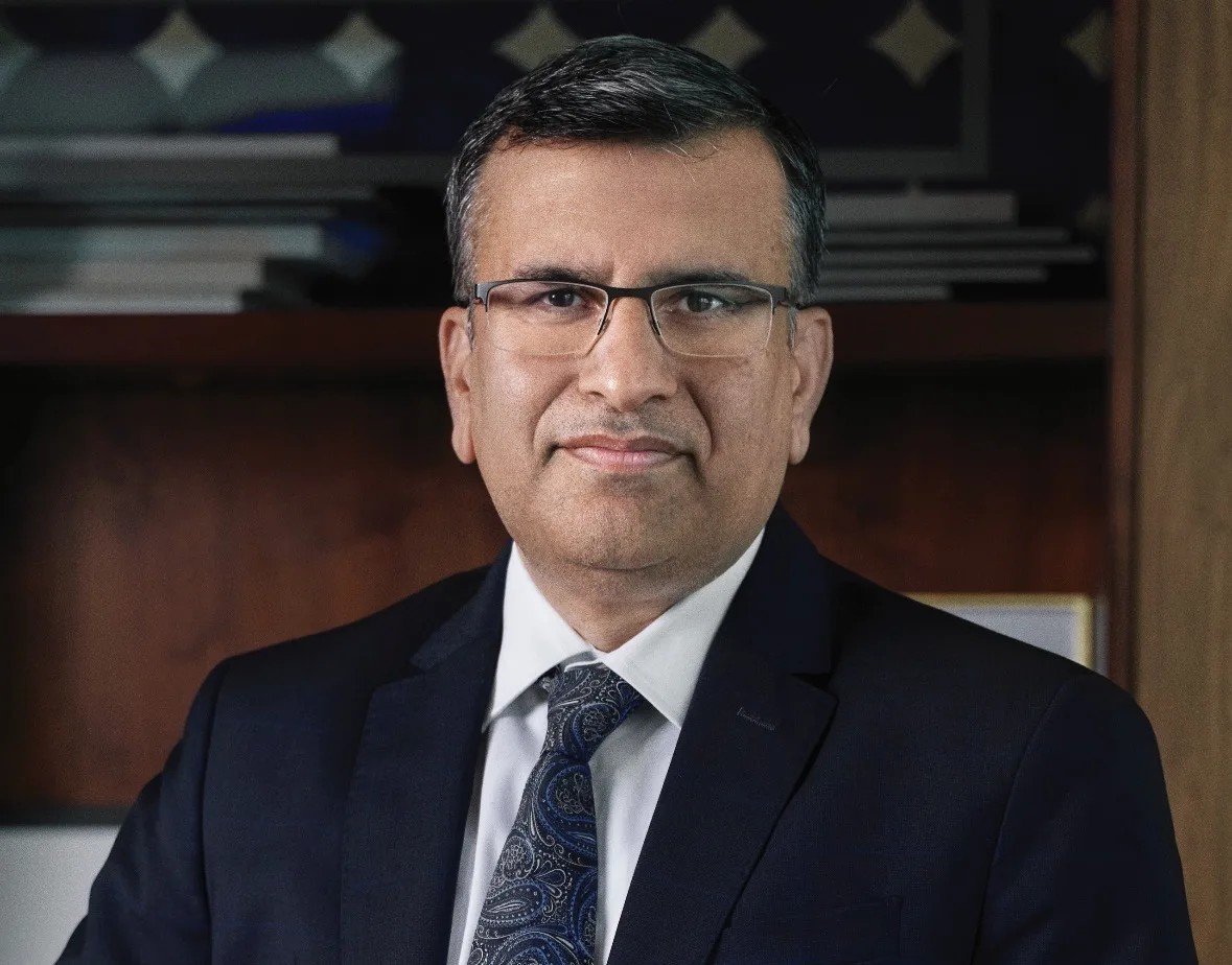 Portrait of a man in glasses, a dark suit and a patterned tie standing in front of a bookshelf