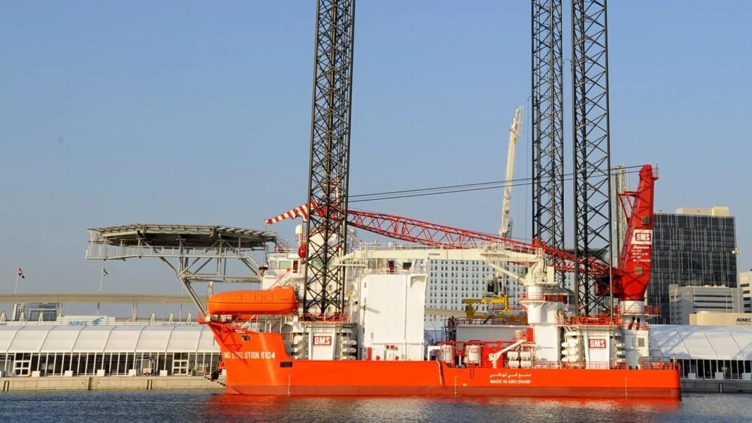 Orange jack-up support vessel with tall lattice legs, a crane and a helideck moored at a quayside beside city towers