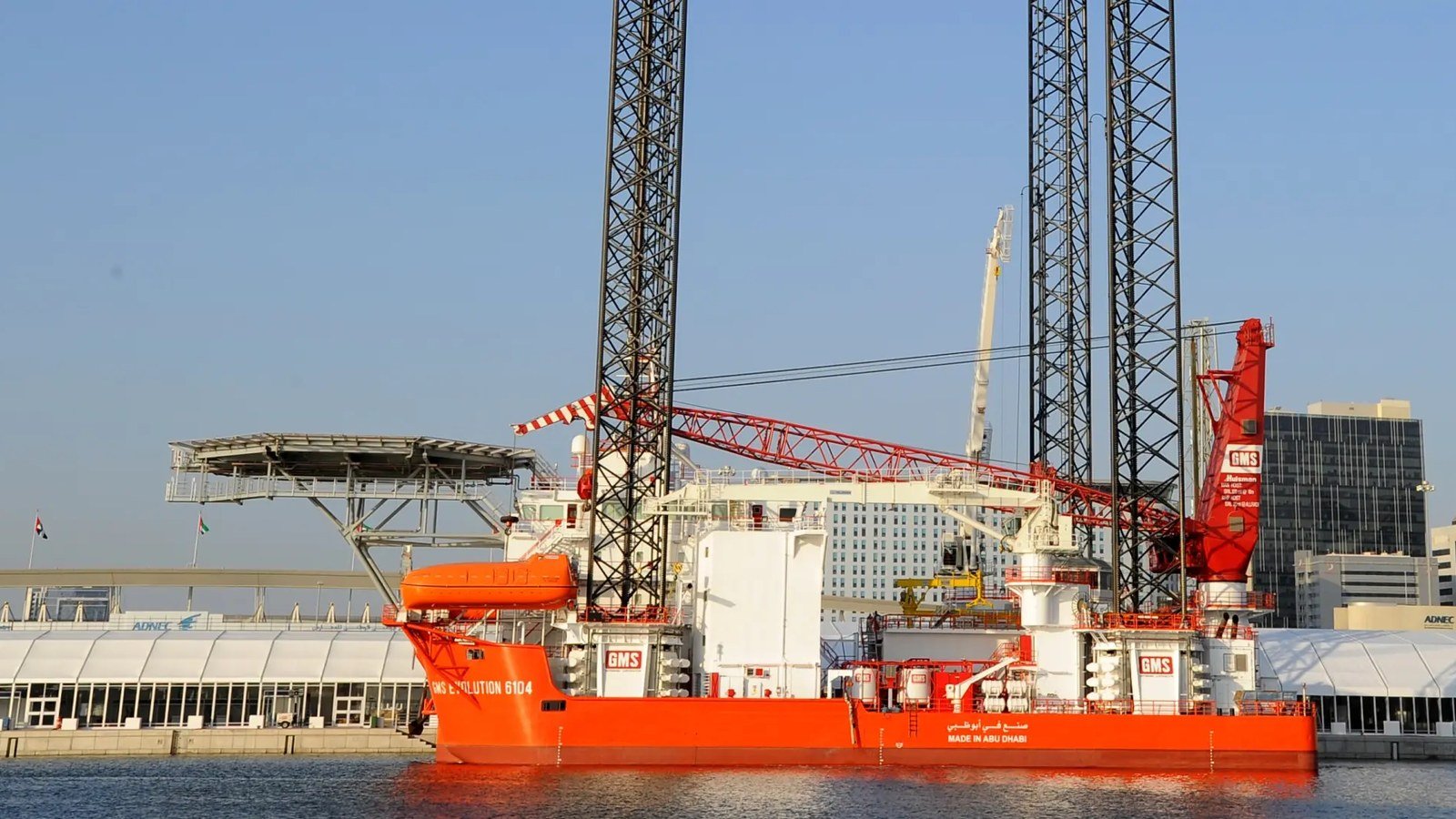 Orange jack-up support vessel with tall lattice legs, a crane and a helideck moored at a quayside beside city towers