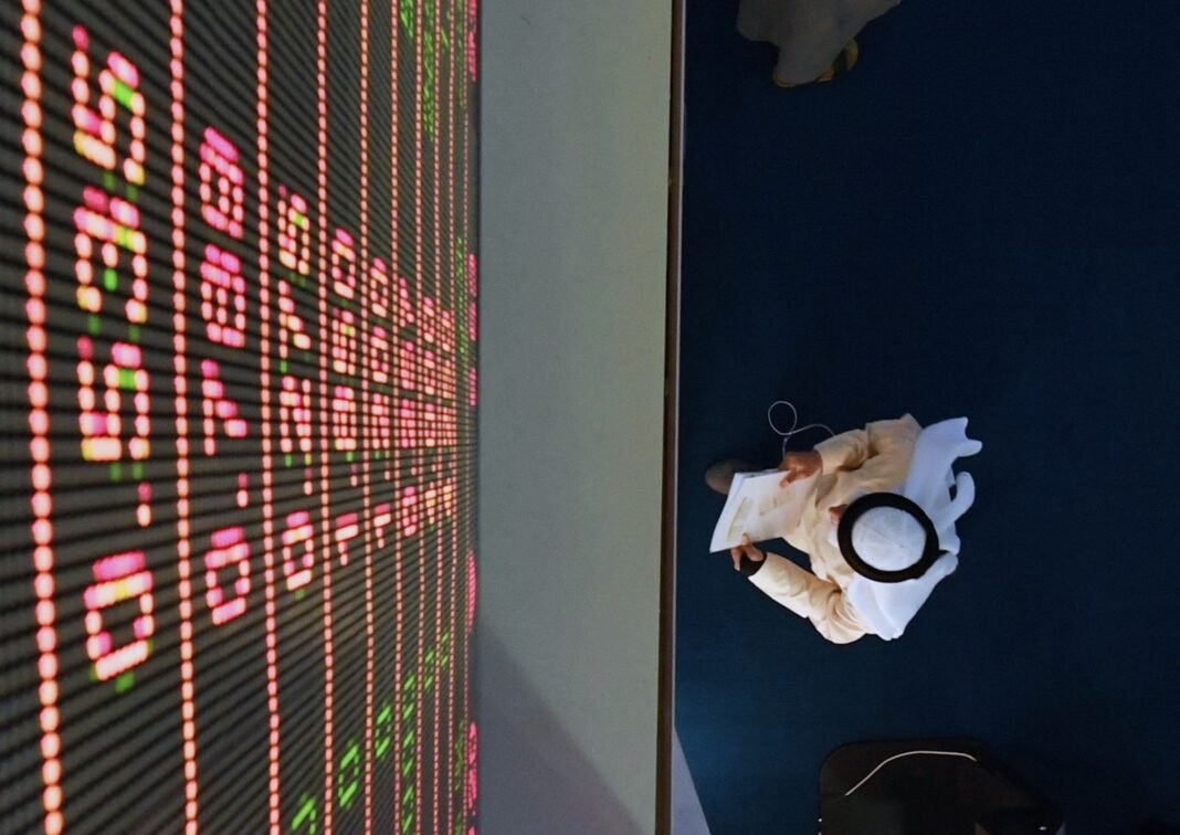 Overhead view of an investor in a white thobe and ghutra walking past an illuminated share price board