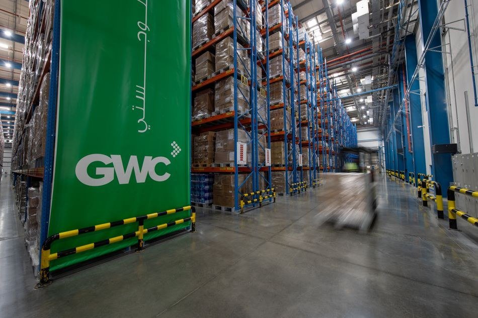 Tall blue and orange pallet racks stacked with boxes line an aisle inside a large warehouse with a green GWC banner